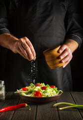 The chef sprinkles salt to salad of fresh vegetables on wooden table. Preparing healthy food on the kitchen