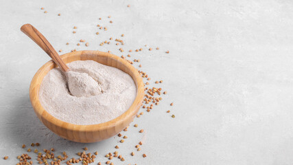 Buckwheat flour in a wooden bowl with wooden spoon and raw green buckwheat grain on light grey...
