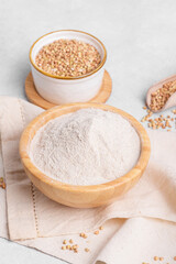 Buckwheat flour in a wooden bowl and raw green buckwheat grain with wooden scoop on light grey table stone background, close up. Alternative flour, gluten free flour, healthy nutrition