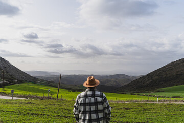 Chico grande con camisa y sombrero frente a paisaje montañoso levantando los brazos y agarrando...