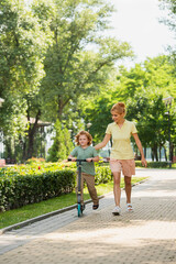 full length view of woman in summer clothes walking near son riding kick scooter in park.