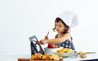 Adorable cute Caucasian little girl using tablet for online learning, cooking in the kitchen alone...