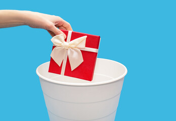 A man throws a gift box into a trash can on a blue background. A human hand holds a red gift box and throws it into a white trash can. Didn't like gift or junk gift concept with free space for text - obrazy, fototapety, plakaty