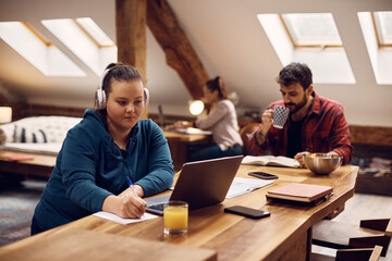 Young woman e-learns on laptop while studying with her roommates in common living room.