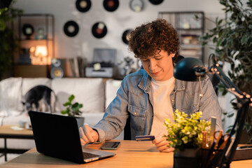 A young man is sitting at his desk in a room next to him with a laptop in his hand holding a payment card. Behind him on the couch lies a dog that is playing with a toy.