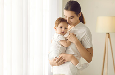 Happy mother together with her child at home. Beautiful young mom standing in a cozy room and holding her cute, sweet baby boy or girl in her arms. Family, love, care, motherhood, and nurture concepts
