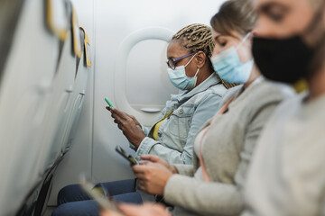 Multiracial people sitting inside aircraft using mobile phone while wearing safety face masks for...