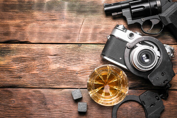 Whiskey in the drinking glass, gun, photo camera and handcuffs on the flat lay detective desk background with copy space.