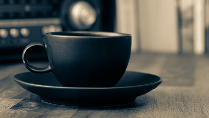 A vintage radio, a black coffee cup and saucer on a brown wooden table, with books in the background, BW vintage look	