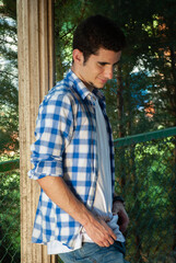 Young man smiling looking at the ground while leaning on a column and surrounded by green garden plants