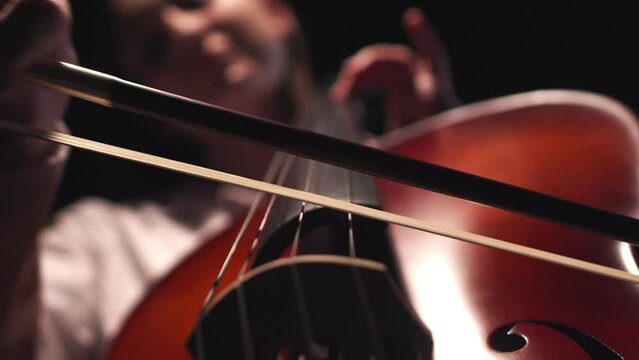 Musician leads bow along strings of cello playing classical music, closeup, front view. Musician performs playing classical melody on cello