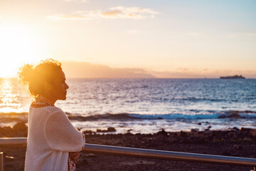 Side view of young adult woman admiring and enjoying golden sunset at the beach looking at the sea. Female people and nature outdoor leisure activity. Thoughtful lady and travel life