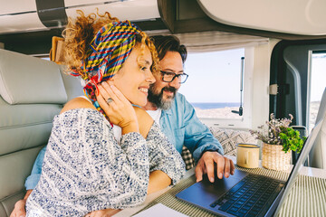 Adult man and woman using together a laptop inside a modern camper van. Couple in digital nomad...