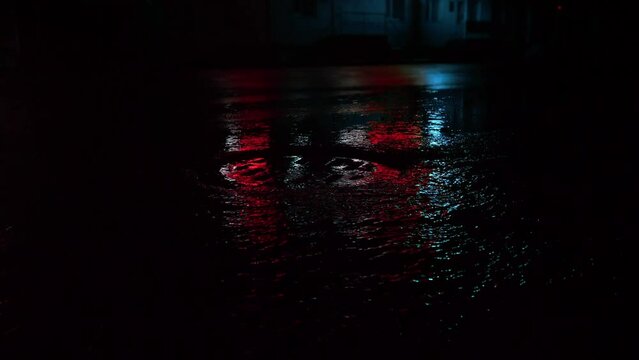 Shimmering water flows through the grate into the storm drain,blood red and blue neon lights are reflected on the wet road during the rain on dark night, copy space