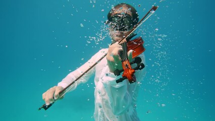 young man figure in underwater sunrays playing violin, boy relaxing and swimming bow along strings. Dolphins playing in the blue water of Red sea. Underwater shot of wild dolphin taking breath
