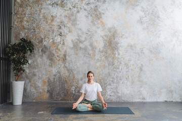 Ardha Padmasana girl sits in lotus position practicing meditation, asana semi-lotos sitting on the floor, yoga mat, spacious gym for training, hands chinmudra - Powered by Adobe