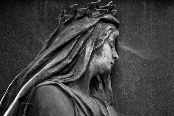 Partial view of an old metal sculpture which shows a grieving, looking to the side, woman with a...