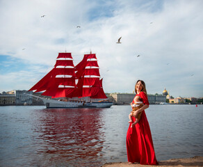 young mother with a newborn baby in a long red dress sees off a ship with scarlet sails on the embankment on a clear summer day - 495983447