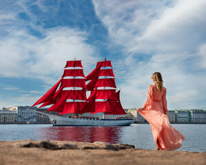young girl in a beautiful long dress meets a ship with scarlet sails on the embankment on a clear summer day - 495983448