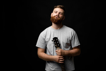 Portraite of handsome brunette bearded man musician, guitarist standing and holding a acoustic guitar in a hand and looks in a camera on a black background studio. Ready to play a music.