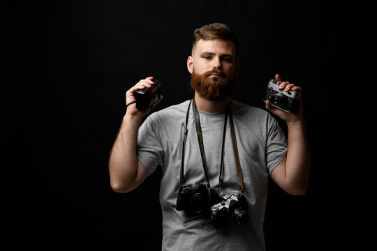 Professional handsome bearded male photographer with bunch of vintage photo cameras in photo studio, isolated on black background.