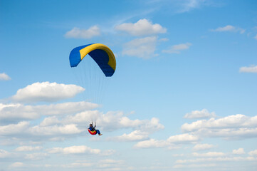 Alone blue and yellow paraglider flying in the blue sky against the background of clouds. Paragliding in the sky on a sunny day.