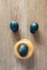 Three painted blue Easter eggs, one in a yellow straw nest on a brown background with natural wood, vertical arrangement, flat lay