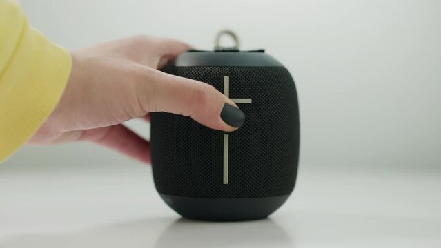 Close-up black portable speaker with female hand pressing plus sign adding volume. Unrecognizable Caucasian young woman listening to music enjoying hobby at white background
