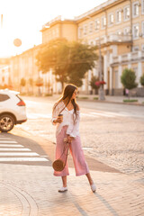 long-haired girl in glasses with phone, coffee and bag walks around the city at sunset