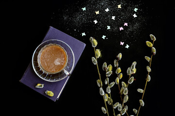 Time for a cup of hot coffee: a cup of coffee stands on a book, next to it is a bouquet of flowering willow, two heart-shaped sugar cubes, a dark background, top view