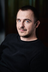 Obraz premium Portrait of a guy in a black jacket, in a dark room. Sly looks into the camera. Blue eyes