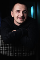 Fototapeta premium Portrait of a guy in a black jacket, in a dark room on a chair. He crossed his arms in front of him and smiled. Blue eyes.
