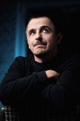 Obraz premium Portrait of a pensive guy in a black jacket, in a dark room on a chair. He crossed his arms in front of him and looked away. Blue eyes