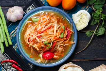 Spicy papaya salad served in a blue dish with sticky rice in a bucket and long beans, dried chilli and vegetables.