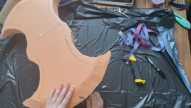 Person's hands cut with knife styrofoam form of Celtic axe. Hatchet for festival or performance. Cosplayer workshop. Handicrafts and diy. Garbage bag on a wooden table.