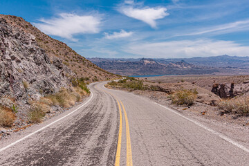Nelson, Nevada, USA - May 21, 2011: Gray asphalt route 165 leads to Colorado River blue water under blue cloudscape, while cutting through dry desert mountainous landscape.