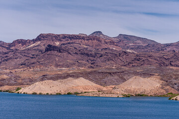 Fototapeta na wymiar Nelson, Nevada, USA - May 21, 2011: Blue Colorado River up front with Arizona shore line and its dry rocky hills and mountains behind under light blue cloudscape. Green plants add color.