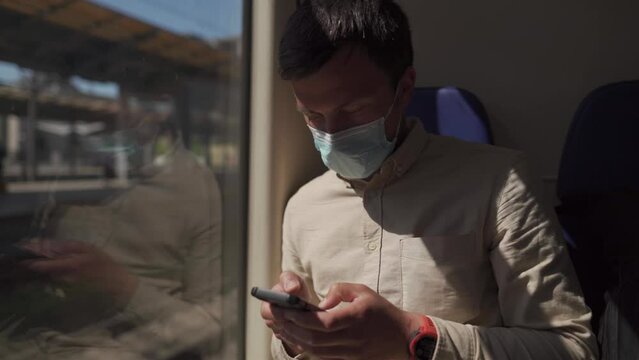 Caucasian man travels in railroad car in face mask and using smartphone, during coronavirus pandemic. Theme of travel and technology. Masked passenger traveling on train surfing Internet on phone