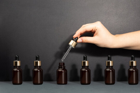 A female hand holds a dropper for applying serum in glass brown bottle. Opened and closed jars of a cosmetic skin care product on a black background. Pattern of beauty products packaging. Skin care
