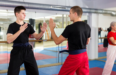 Fototapeta na wymiar Focused young guy in black sportswear sparring with experienced martial arts trainer during training in gym