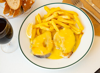 Stewed hake fish with melted cheese, french fries and lemon on a plate in a restaurant