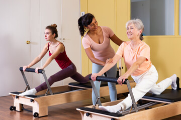 Fototapeta na wymiar Aged woman doing pilates on reformer in fitness studio with Hispanic female personal trainer controlling movements