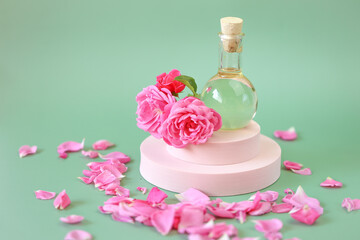 Rose essential oil in a glass bottle in rose flowers in rose petals on a pink podium on a green...