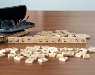 ugly duckling word or concept represented by wooden letter tiles on a wooden table with glasses and a book - obrazy, fototapety, plakaty