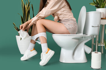 Young woman with paper sitting on toilet bowl against green background