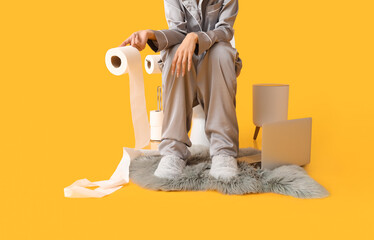 Young woman in pajamas and with paper sitting on toilet bowl against color background