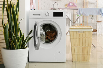 Modern washing machine with laundry, basket and plant in bathroom