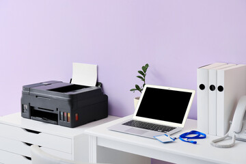 Workplace with modern laptop and chest of drawers with printer near color wall