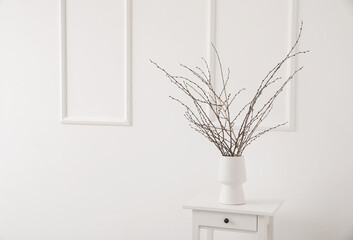 Vase with tree branches on white table near light wall