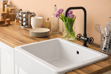 Fototapeta na wymiar Ceramic sink, kitchen utensils and vase with tulips on counter near beige wall